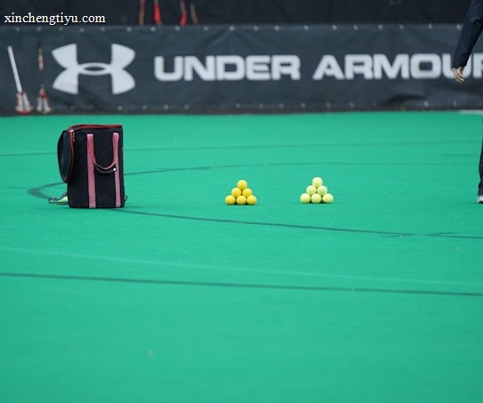 A sports field features a black and pink sports bag, two pyramids of yellow balls, and a portion of a person in dark athletic gear. The background displays a banner with the Under Armour logo.