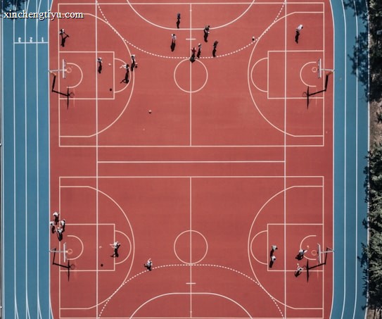 An aerial view of a large, vibrant sports complex featuring a red basketball court with multiple hoops and a surrounding blue running track. Several people are scattered across the area, engaged in various activities.